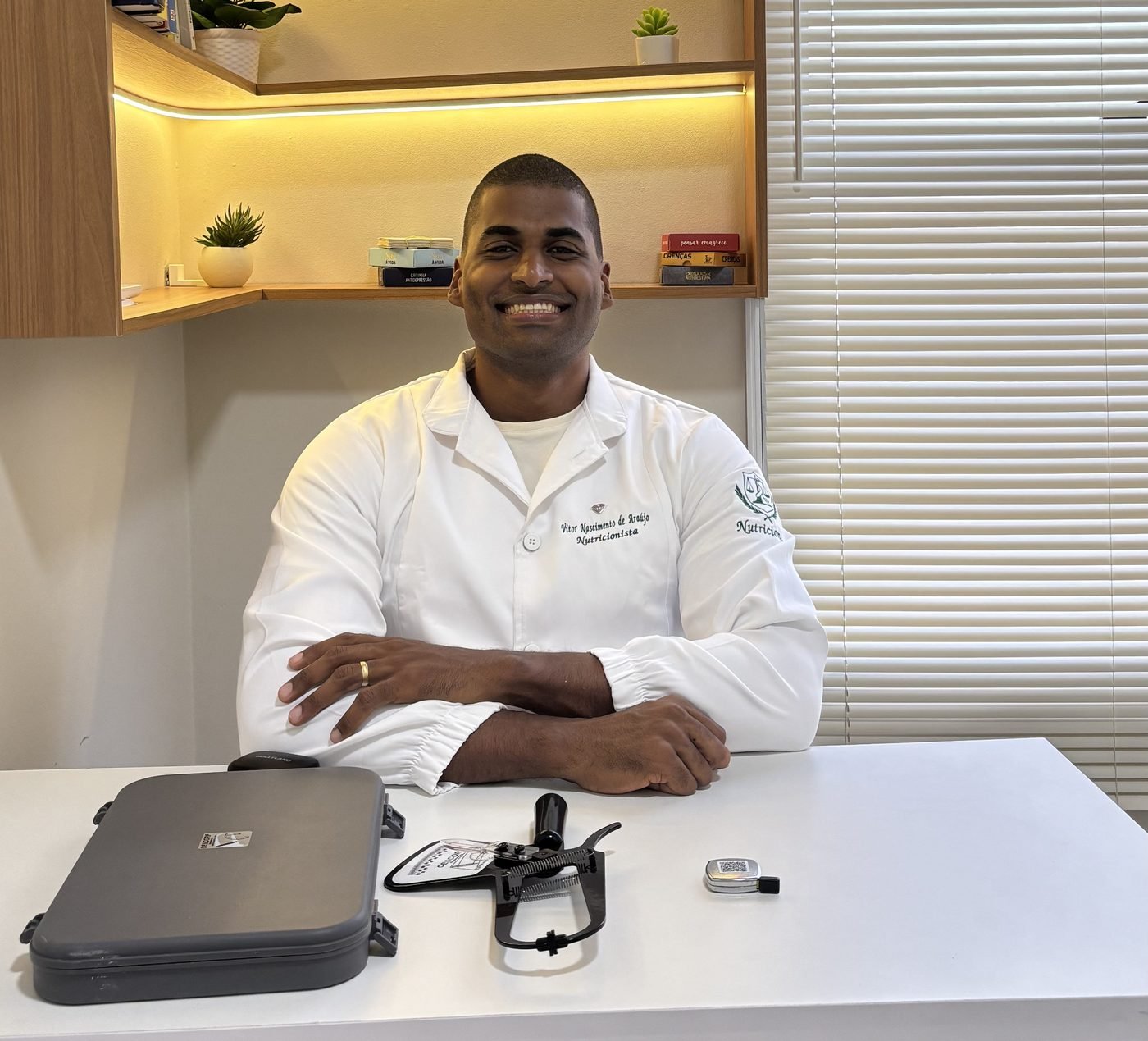 Vitor Nascimento, nutricionista, sorrindo à mesa do consultório em Manaus, com o adipômetro à frente